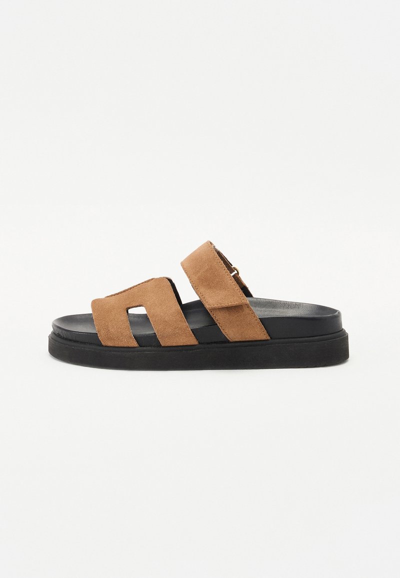 Bruine suède slide sandalen met twee brede banden, een zwarte rubberen zool en een glad, plat ontwerp. Heeft minimale accenten en een open teen.