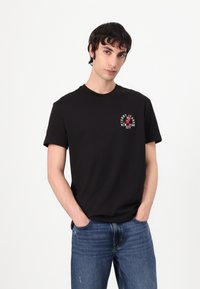 Black cotton t-shirt with a round neck, short sleeves, and a embroidered logo depicting a red figure and text. Worn with blue jeans.