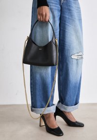 Sac en cuir noir avec une forme courbée, une courte poignée et une bandoulière en chaîne dorée. Porté avec un jean bleu déchiré et des escarpins noirs.