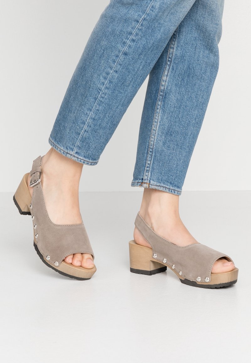 Chaussures grey en daim à bout ouvert avec une bride arrière, talon en bois et clous argentés sur le côté, accompagnées de jeans en denim bleu.