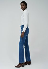 Jeans acampanados azules con una cintura alta, combinados con una blusa blanca de manga larga. Botas marrones de tacón con detalles metálicos.