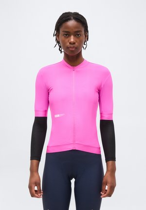 Žena oblečená v jasne ružovom cyklistickom tričku s čiernymi rukávmi a tmavých textúrovaných cyklistických kraťasoch na jednoduchom pozadí.