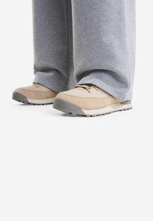 Beige og grå udendørs sko brugt med løse grå joggingbukser, vist fra siden og front på en hvid baggrund.