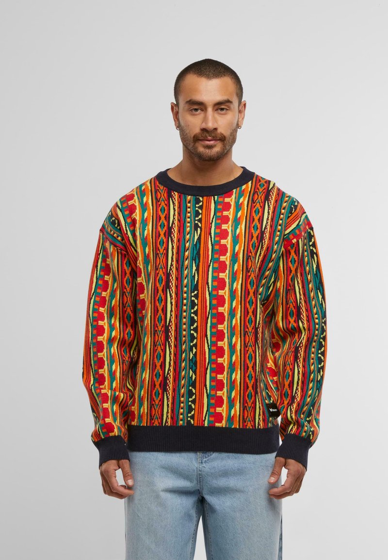 Multicolor trui met gedurfde geometrische patronen in rood, groen, geel en zwart, met een geribbelde marineblauwe kraag en boorden.