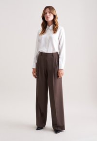 Witte, langmouwige blouse met subtiele strepen, gecombineerd met hooggesneden, brede bruine broeken en zwarte puntige schoenen.
