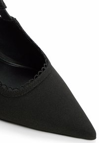 Zapato negro de punta con una parte superior de tela texturada. Presenta un acabado con borde festoneado y un diseño de lado abierto para mayor transpirabilidad.