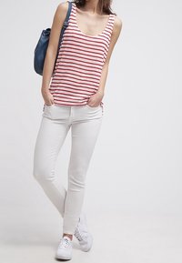 Mladá žena stojící s rukama v kapsách, na sobě má červenobílé pruhované tílko, bílé skinny džíny, bílé tenisky a nosí modrý batoh.