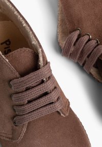 Le scarpe in camoscio marrone presentano sei lacci intrecciati e un morbido rivestimento interno. Il design include un dettaglio cucito sulla punta.