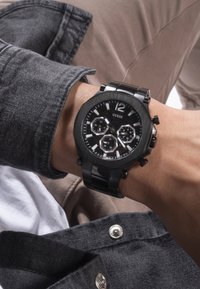 Schwarze Guess Armbanduhr mit silbernen Akzenten, großem rundem Zifferblatt, Chronographen-Dials und glänzendem Armband. Am Handgelenk mit einer grauen Jacke getragen.