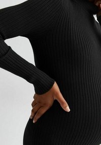 Person som bär en svart ribbad långärmad tröja, med ena handen vilande på sin nedre rygg och den andra handen som rör vid sin nacke.