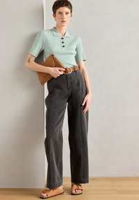 Polo shirt a righe verde chiaro con bottoni, jeans a gamba larga grigio scuro, cintura in pelle marrone, pochette marrone e sandali color sabbia con accenti metallici.