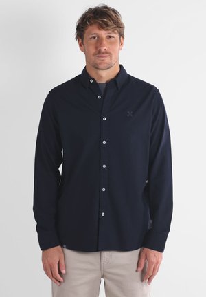 Chemise bleu marine à boutons avec une coupe ajustée, manches longues et boutons blancs. Présente une broderie discrète sur le côté gauche de la poitrine. Tissu léger.