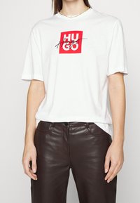 Camiseta blanca de algodón con un logo cuadrado rojo que presenta "HUGO" en cursiva negra. Llevada con pantalones de cuero marrón, mostrando un ajuste relajado.