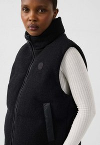 Donna che indossa un gilet in pile nero con zip sopra una maglia bianca a maniche lunghe a coste, rivolta leggermente a sinistra con un'espressione neutra.