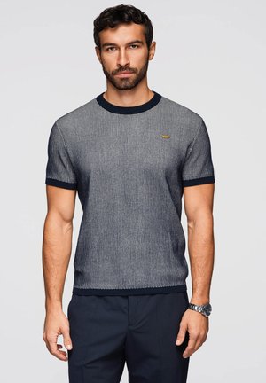 Hombre con cabello corto y oscuro y barba, vistiendo una camisa de punto de manga corta y ajustada de rayas marinas y pantalones oscuros, de pie frente a un fondo liso.