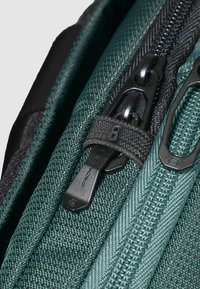 Dettaglio dello zaino con tessuto verde scuro testurizzato, cerniere nere e cursore con etichetta. Primo piano sulle cuciture e sui componenti hardware.