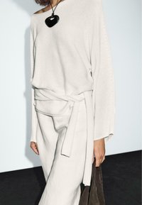 Light gray, knitted sweater with oversized sleeves and a tie belt, paired with light gray, loose-fitting pants. Black heart-shaped pendant necklace.