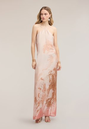 Junge Frau trägt ein langes, halterneck pink-braun gemustertes Kleid mit Riemchensandalen, steht vor einem schlichten Hintergrund.