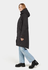 Schwarzer langer Mantel mit Kapuze, ausgestattet mit zwei Seitentaschen und knöpfbaren Manschetten. Kombiniert mit hellblauen Jeans und schwarzen klobigen Stiefeletten.