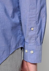 Helles blaues Langarmhemd aus strukturiertem Stoff. Verfügt über zwei Knöpfe am Ärmel und ein schräges Taschen-Detail am Ärmel.