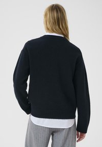 Navy blue knitted sweater with a relaxed fit, ribbed cuffs and hem. Worn over a white collared shirt, showing textured fabric and minimal design.