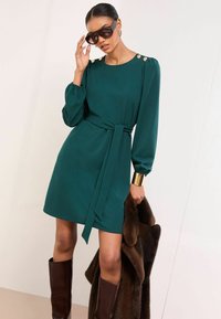 Zelené šaty s opaskom a dlhými rukávmi, zlatými gombíkmi na ramenách a hladkou textúrou, doplnené hnedými čižmami po kolená a hnedým kožušinovým kabátom.