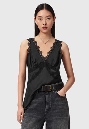 Jonge vrouw met lang donker haar draagt een zwarte mouwloze top met kanten details, een donkerpaarse high-waisted spijkerbroek, een zwarte riem en een zilveren ketting.