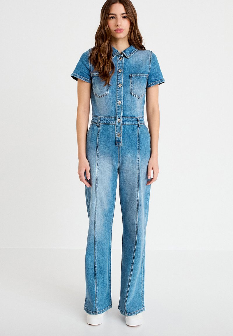 Mujer de pie, mirando hacia adelante, vistiendo un mono de mezclilla azul de manga corta con botones y zapatos blancos sobre un fondo blanco.