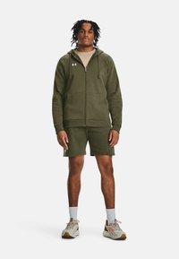 Olivgrüner Hoodie mit Vorderreißverschluss und Taschen, kombiniert mit passenden Shorts. Sportliche Schuhe mit hellen und dunklen Akzenten.