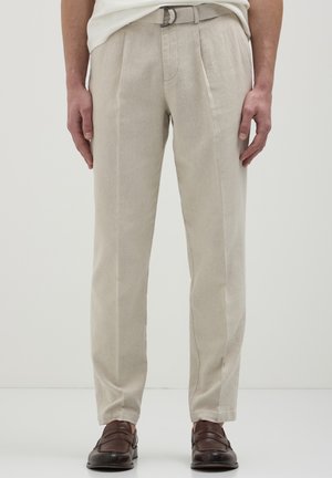 Mann trägt beige maßgeschneiderte Hose mit einem Gürtel, kombiniert mit dunkelbraunen Leder-Leder-Schuhen, stehend vor einem schlichten Hintergrund.