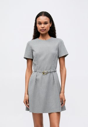 Eine Frau trägt ein kurzärmliges schwarzes und weißes Hahnentrittkleid mit passendem Gürtel und goldener Schnalle und steht vor einem schlichten hellen Hintergrund.