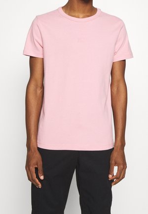 Person wearing a light pink short-sleeve crew neck t-shirt and black pants, standing with arms relaxed by sides.