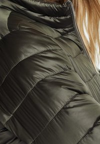 Chaqueta de plumas de color verde oliva con un diseño acolchado y diferentes texturas. El alto cuello y el cierre de cremallera destacan el material cálido y aislante.