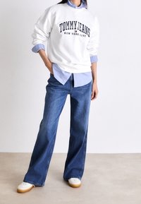 Sudadera blanca con la impresión "TOMMY JEANS", superpuesta sobre una camisa de botones azul claro. Jeans azules acampanados y zapatillas blancas con suelas en tono beige.