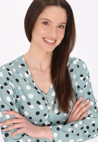 Jonge vrouw met lang, bruin haar die glimlacht, gekleed in een lichtgroene blouse met zwart-witte stippen, met de armen over elkaar.