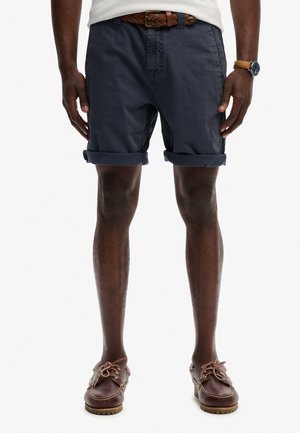 Mann trägt marineblaue, hochgekrempelte Shorts, geflochtener brauner Gürtel, weißes Hemd, braune Lederuhr und braune Leder-Schnürschuhe.
