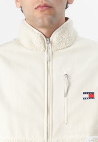 Kremfarget jakke med fleece-kant, glidelås foran, høy krage og brystlomme med et blått, rødt og hvitt logo-merke.