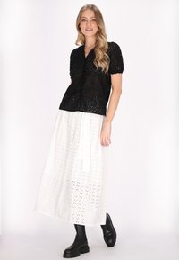 Blouse noire en dentelle avec des boutons et des manches bouffantes ; associé à une Jupe blanche en broderie anglaise au design fluide et des bottines noires à la cheville.
