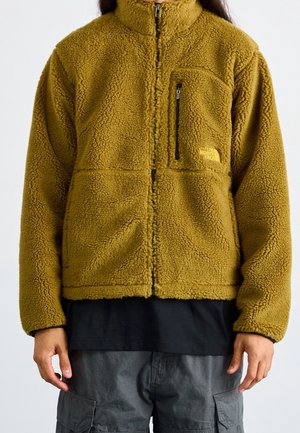 Persoon draagt een olijfgroene fleecejas met ritssluiting aan de voorkant en een ritssluiting op het borstzakje, gecombineerd met een zwart overhemd en grijze cargobroek.