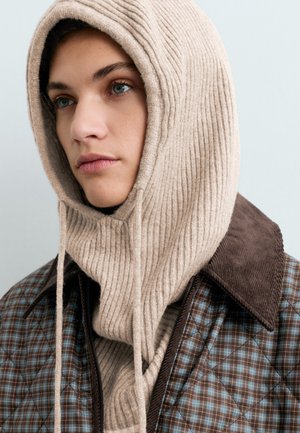 Portrait en gros plan d’une personne portant une capuche beige en maille côtelée et une veste à carreaux bleu-brun avec un col foncé.