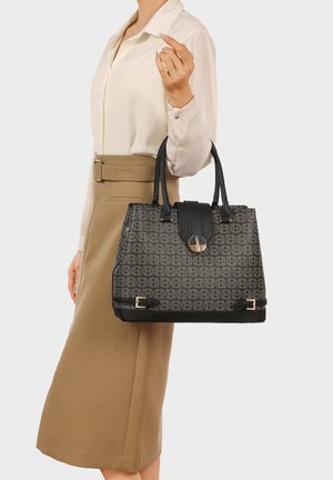 Donna con gonna beige e camicetta bianca che tiene una borsa a motivo grigia e nera con doppi manici e dettagli dorati.