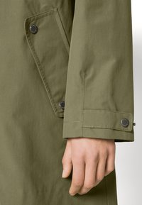 Oliwkowy płaszcz z gładkim, teksturowanym materiałem. Posiada guzik na mankiecie oraz ukośną kieszeń z widocznymi detalami szwów.