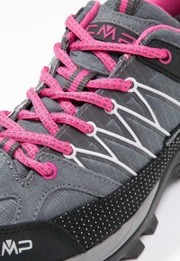 Zapato deportivo gris con cordones rosas, con parte superior texturizada, detalles en blanco y un patrón de puntos negros a lo largo de la suela.