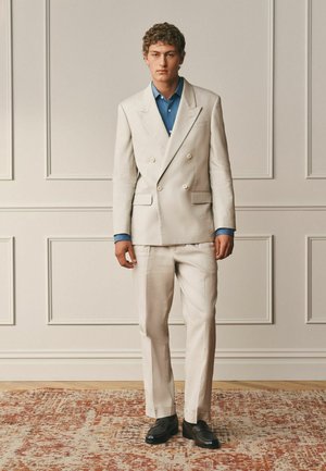 Nuori mies kiharat hiukset, pukeutunut kermaan kaksiriviseen pukuun, siniseen paitaan ja mustiin kenkiin, seisoo kuvioidun maton päällä paneeliseinää vasten.