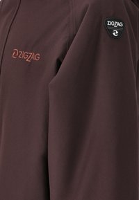 Donker bordeauxkleurige jas met een gladde stofafwerking. Het heeft een zwart embleem met "ZIGZAG" en een close-up van het geborduurde logo op de mouw.