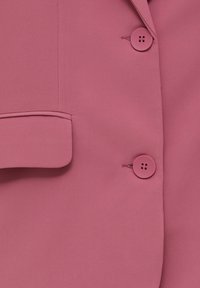 Gros plan sur le devant d'une veste rose montrant deux boutons ronds et une poche à rabat sur un tissu lisse.