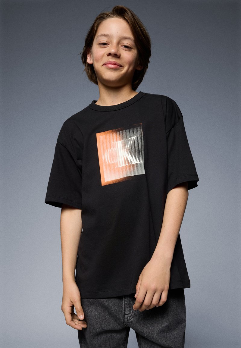 Schwarzes Baumwoll-T-Shirt mit kurzen Ärmeln, das ein zentrales Grafikdesign in Orange und Weiß zeigt, und in lockerem Schnitt. Grauer Hintergrund.