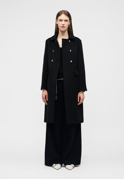 Γυναίκα που φορά μαύρο παλτό διπλού πέτου, μαύρο παντελόνι φαρδιάς γραμμής, ζώνη με αλυσίδα και λευκά μυτερά παπούτσια με αγκράφα, στέκεται μπροστά σε απλό φόντο.