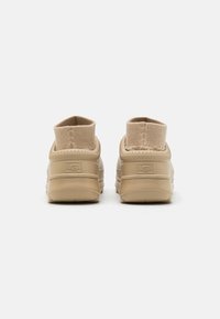 Scarpe slip-on beige con un colletto in maglia testurizzata e suola in gomma. Logo UGG sul tallone, con un design a retro arrotondato.