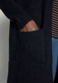 Laivastonsininen neulottu villatakki, jossa on ribatut hihat ja etutasku. Kangas näyttää pehmeältä ja siinä on strukturoitu pinta, kerroksittain raidallisen paidan päällä.